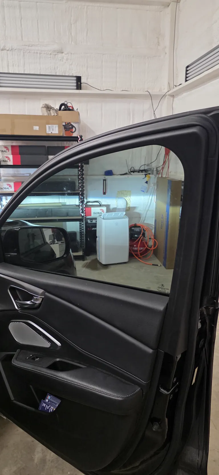Freshly tinted front door window on a black Acura, shop shelves of film visible through the glass