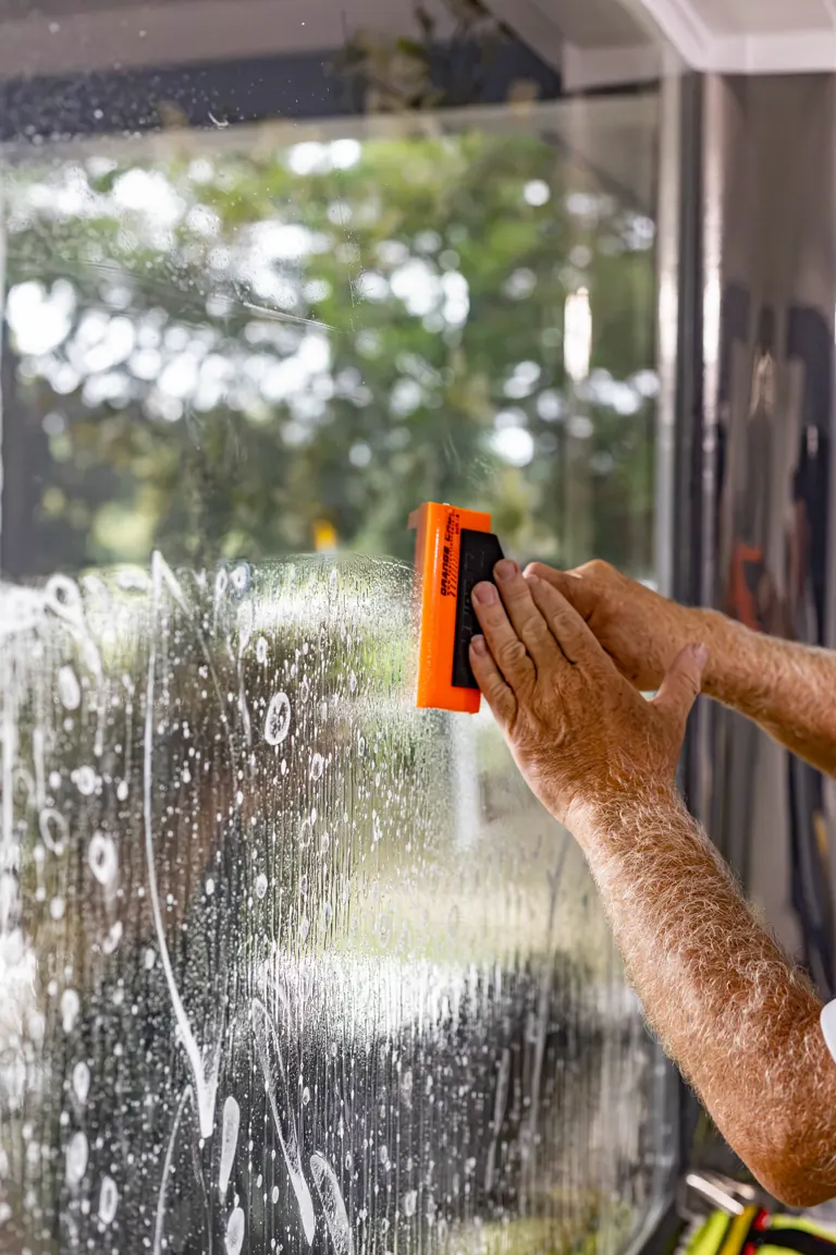Hands drawing a squeegee down wet film on a home window, water beading and running under the blade